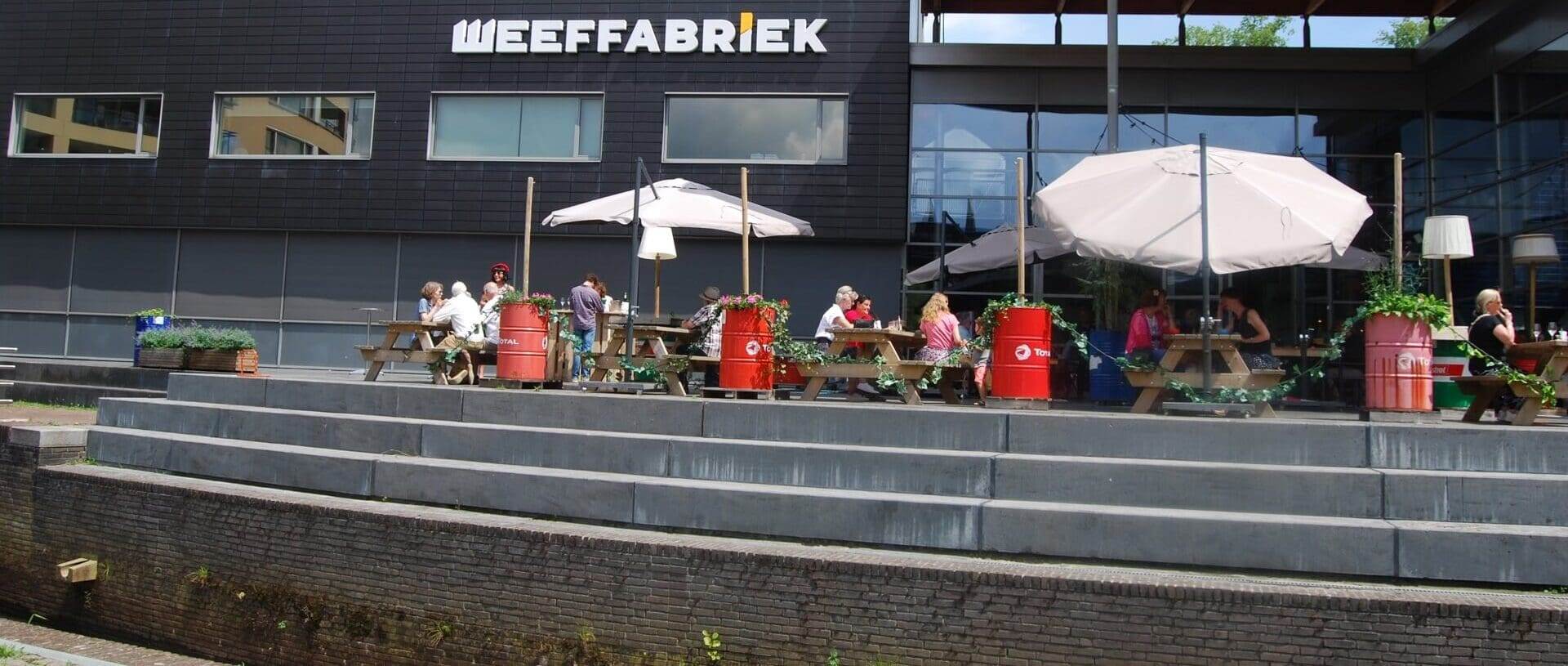 Jongeren genieten op het gezellige buitenterras van de Weeffabriek in Veghel, waar houten picknicktafels en kleurrijke rode tonnen als tafeldecoratie zorgen voor een levendige sfeer. Grote parasols bieden schaduw terwijl vrienden met elkaar kletsen en ontspannen. Op de achtergrond zie je het moderne, zwarte gebouw van de Weeffabriek met grote ramen en het opvallende logo boven de ingang - dé hotspot om samen te komen in Veghel.