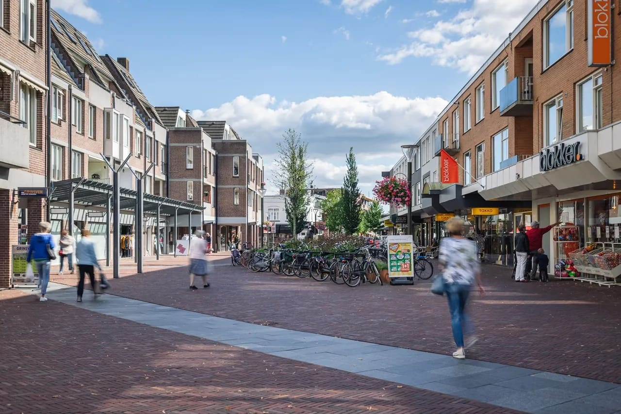 Drukke winkelstraat met moderne bakstenen panden, gezellige winkels, balkons en fietsen. Bloembakken sieren lantaarnpalen.