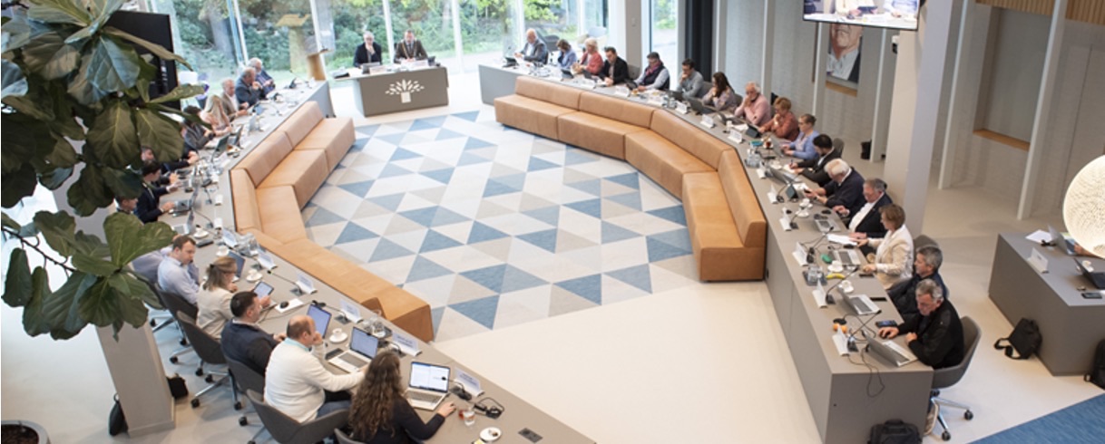 In een moderne vergaderruimte werken mensen actief aan laptops rond een U-vormige tafel, terwijl de spreker centraal zit.