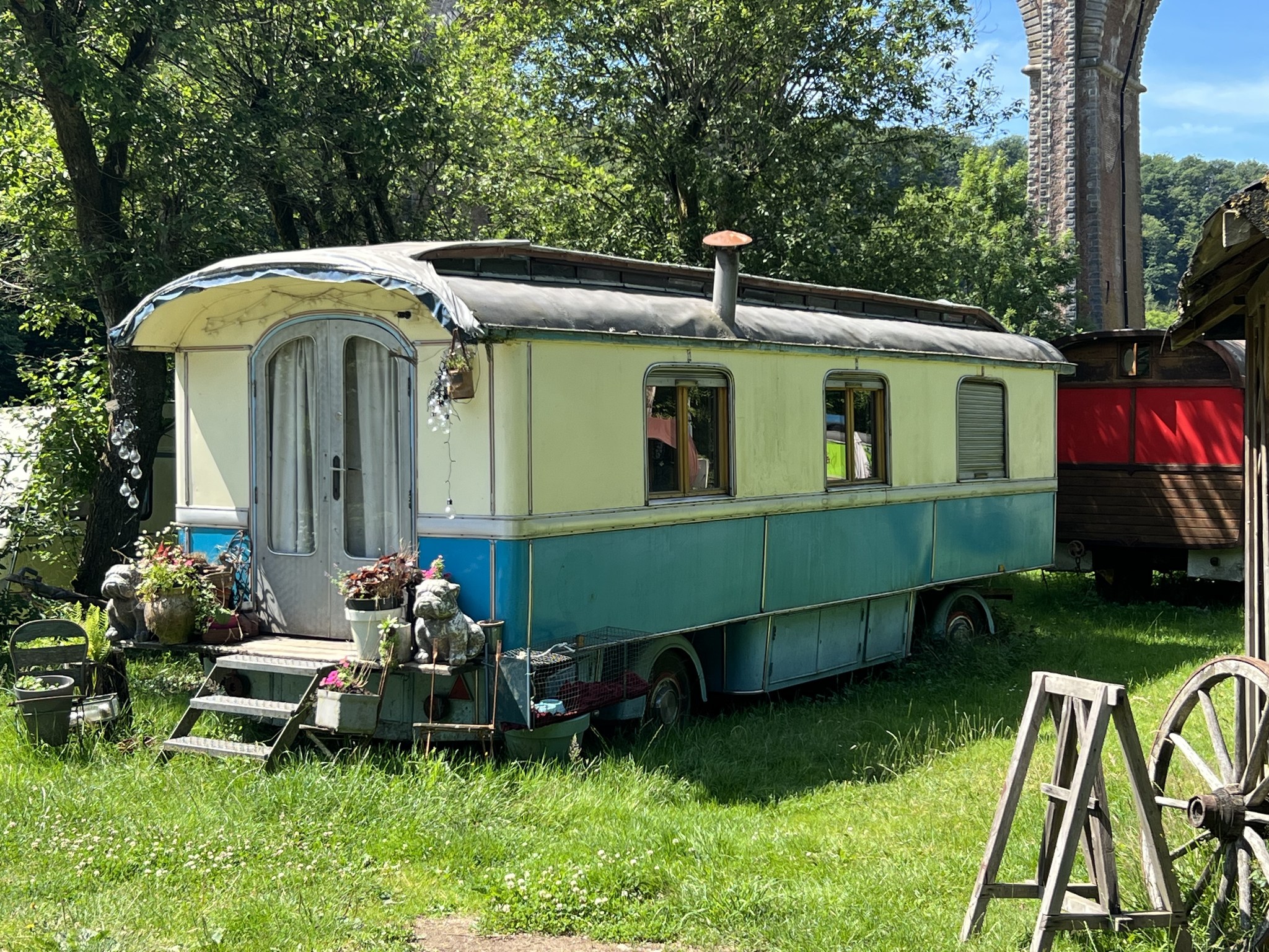 Een retro blauw-rode caravan staat op gras, omringd door planten en tuinaccessoires; op de achtergrond een stenen viaduct.
