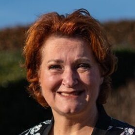 Mariëlle Verdouw-de Vet lacht buiten in de zon, omringd door groene natuur en blauwe lucht, in een bloemenjasje.