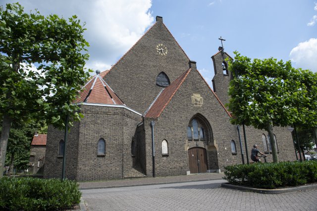 Op het Sint Jozefplein prijkt een bruine bakstenen kerk met steil dak en houten deuren; iemand stopt bij de ingang onder wolkenluchten.