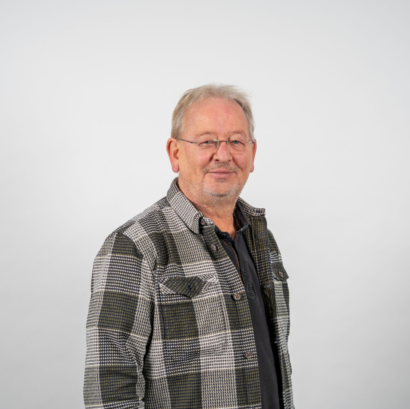 Een vriendelijke oudere man met grijs haar, bril en baard glimlacht in een moderne geruite blouse voor een witte achtergrond.