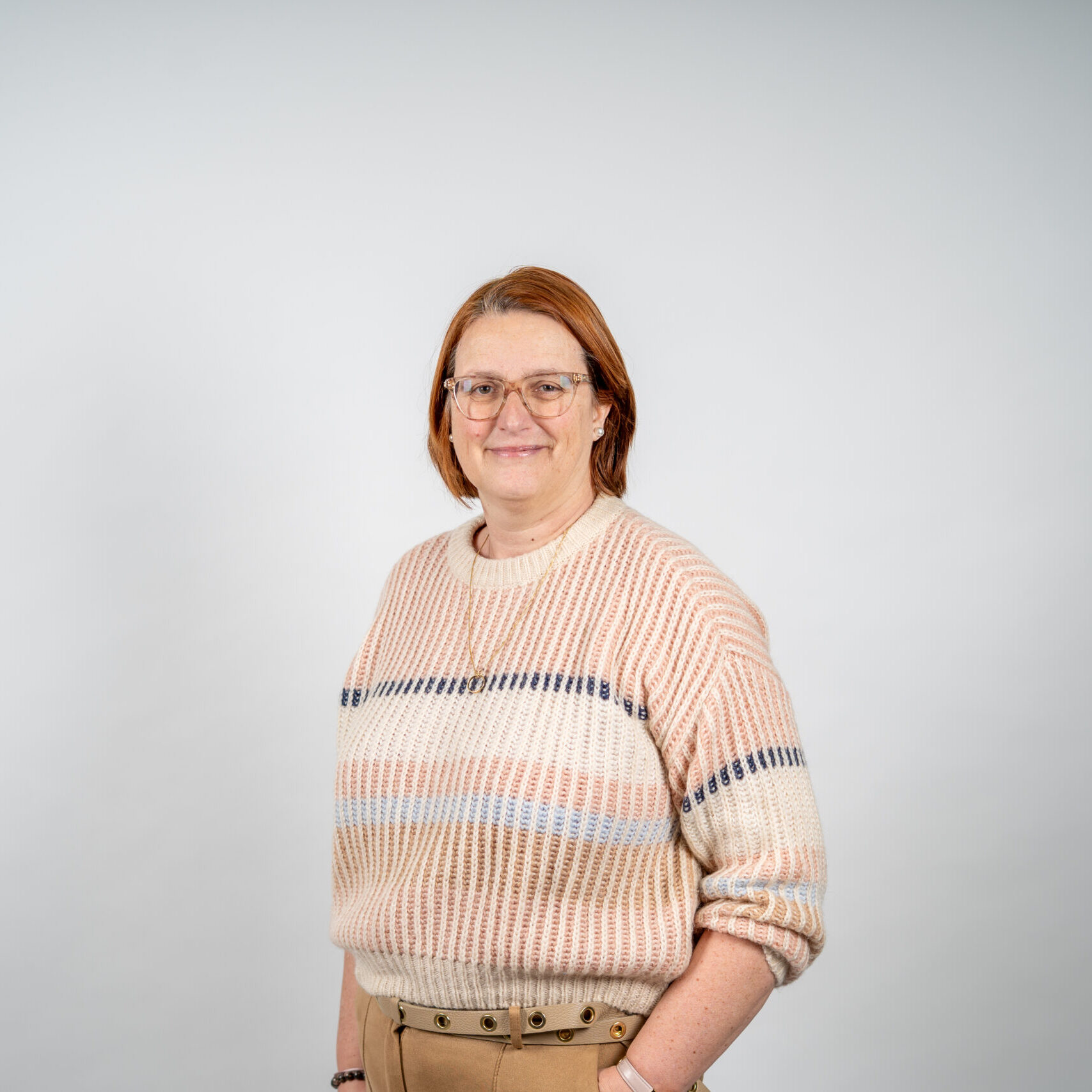 Esther Liefbroer, met kort rossig haar en bril, straalt in een gestreepte trui en beige broek voor een lichte achtergrond.