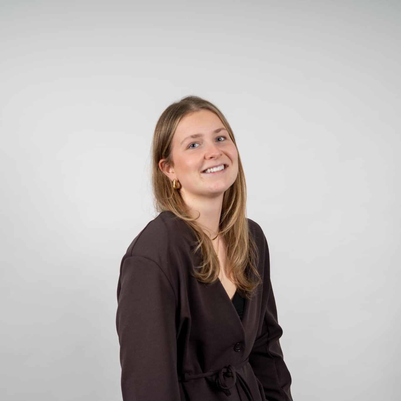 Jonge vrouw met lang lichtbruin haar lacht naar de camera in stijlvolle jas met gouden oorbellen, voor een grijze achtergrond.