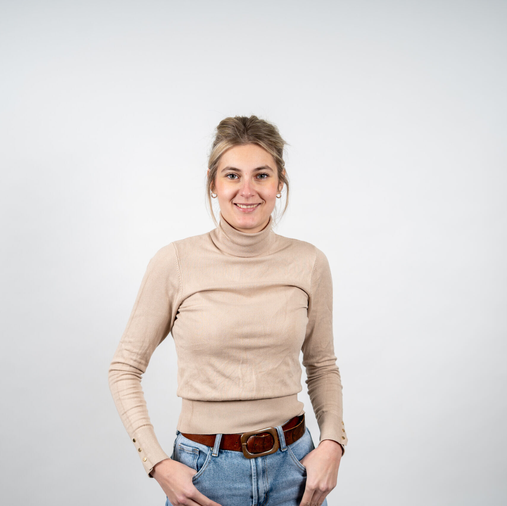 Vrouw met lichtbruine opgestoken haren straalt in beige coltrui, blauwe jeans en bruine riem tegen een witte achtergrond.