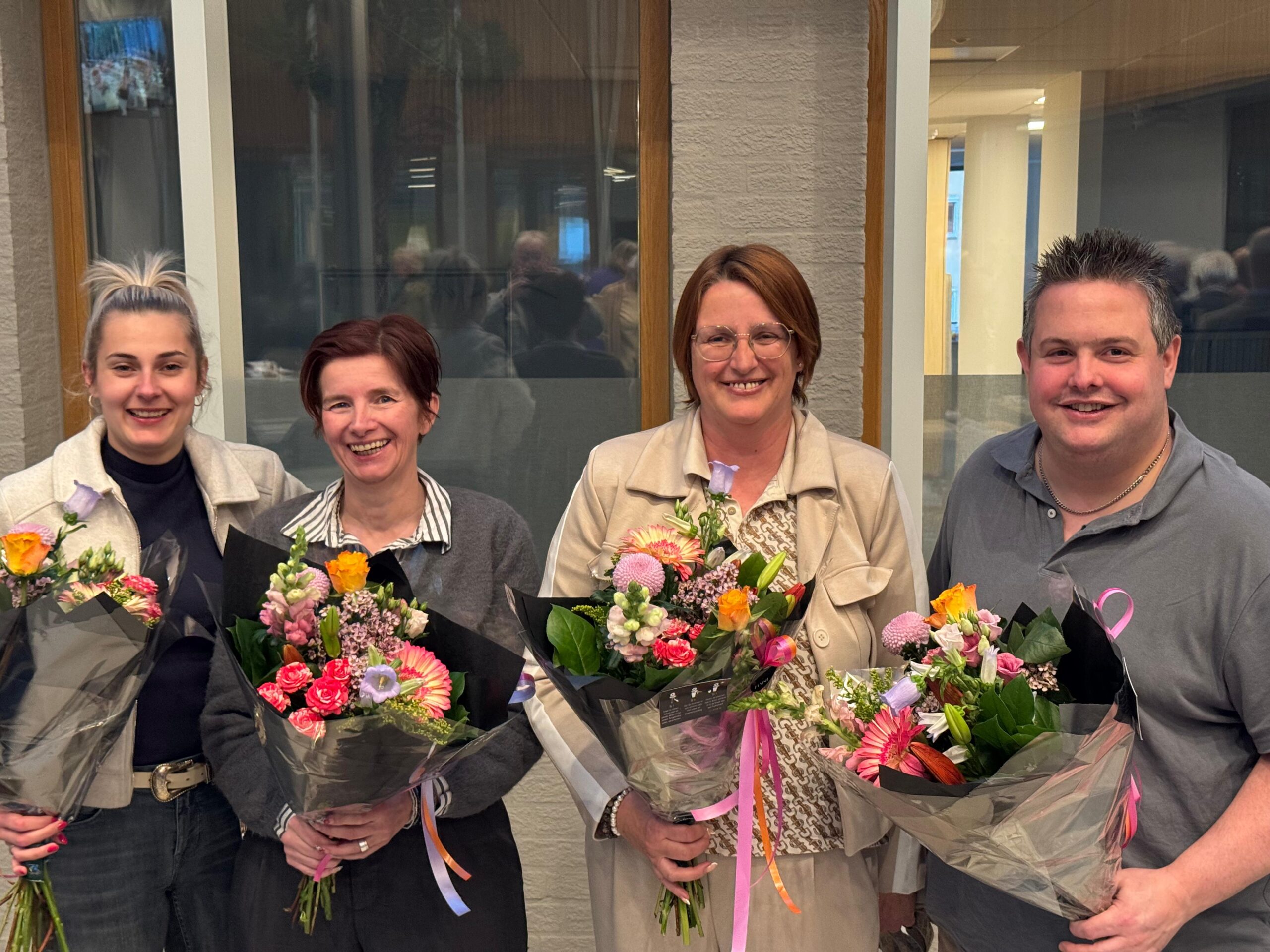 Vier commissieleden van SAMEN Geldrop-Mierlo poseren binnen met bloemen voor grote ramen bij de wisseling van de wacht.